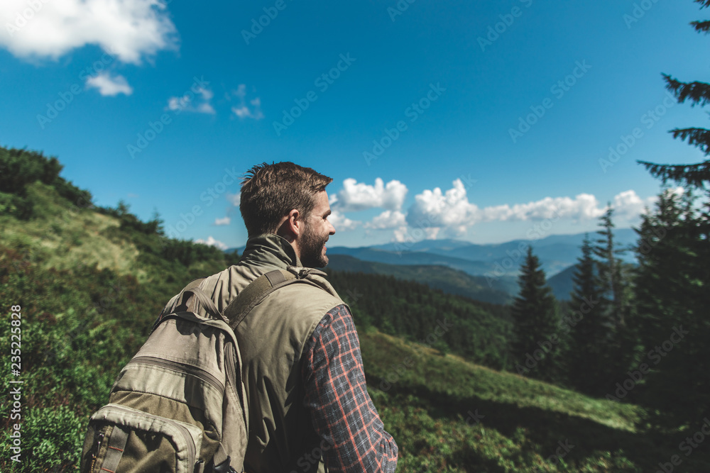 Concept of breathtaking journey and adventure. Waist up back side portrait of smiling man enjoying the nature view while standing on green hill of mountain