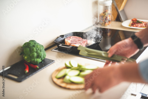 Close up of beef steak with garlic on frying pan. Blurred male hands holding kitchen tongs and cutting board with chopped zucchini