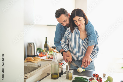 Dinner for two. Waist up portrait of charming young lady chopping onion while bearded man hugging her from behind and showing how to cut properly