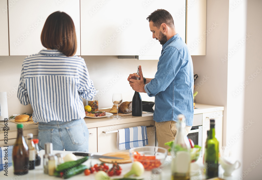 Almost ready. Portrait of bearded man opening bottle of wine young lady standing near food Almost ready. Portrait of bearded man opening bottle of wine young lady standing near food