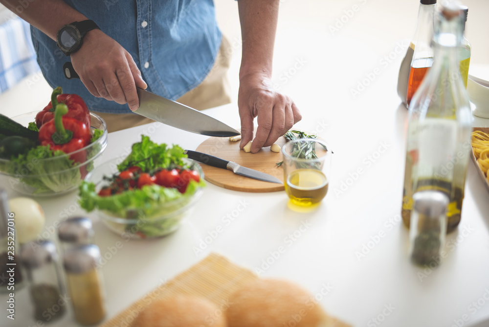 Close up of male hands with knife cutting clove of garlic. Glass of olive oil and bowls with vegetables on white kitchen table