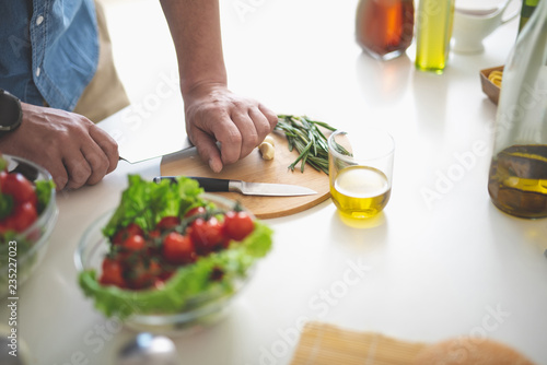 Close up of male hands with knife preparing ingredient for fresh salad. Glass of olive oil and bowl with vegetables on kitchen table