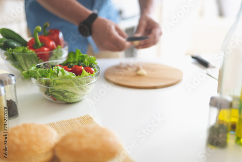 Fototapeta Naklejka Na Ścianę i Meble -  Healthy food. Close up of bowls with tomatoes, lettuce, cucumber and red bell pepper on kitchen table. Male hands with knife on blurred background