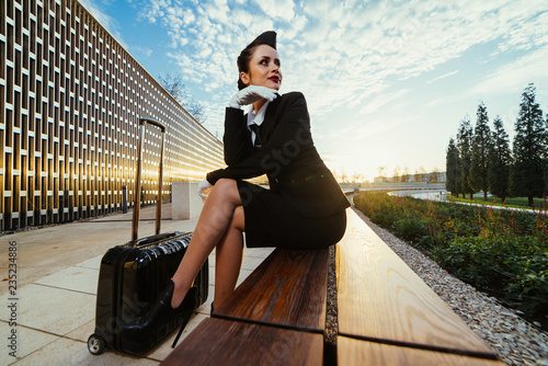 stylish beautiful young woman stewardess in uniform sits on a bench and waits for her flight, with a suitcase photo