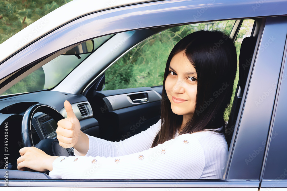 Happy young girl with long dark hair driving a car and gesturing thumb up