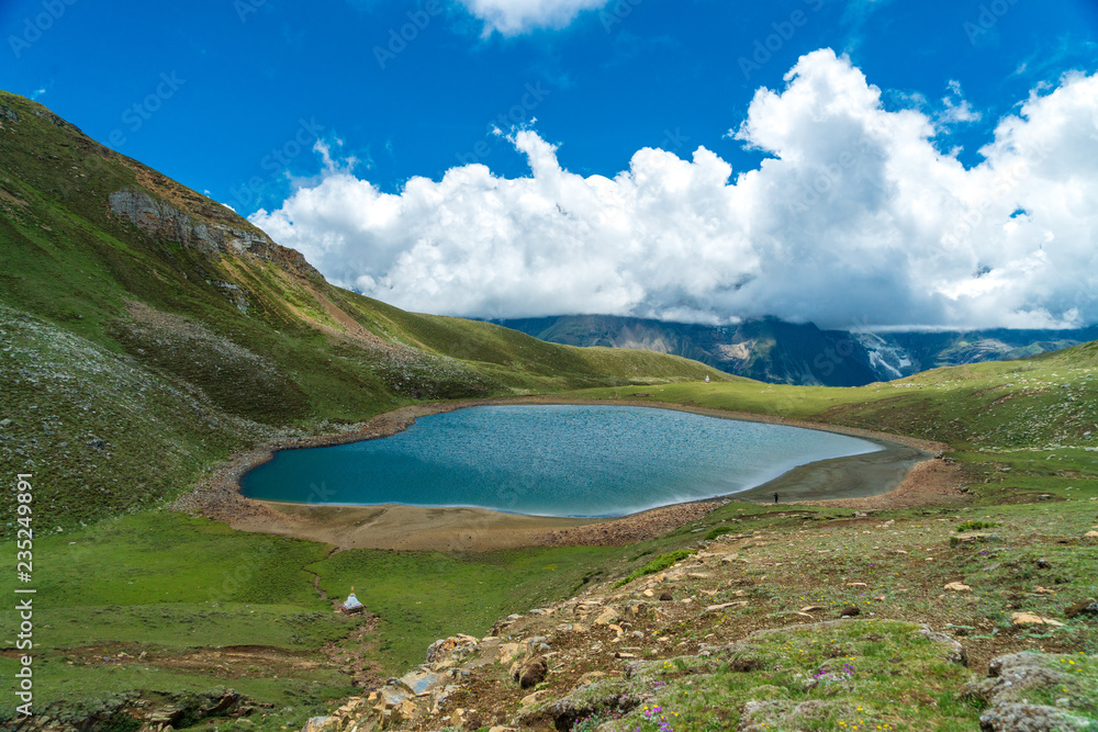 A lone figure stands on the edge of a blue alpine lake with fluffy white clouds floating overhead on a summer day