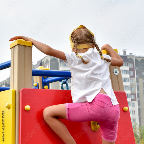 A pigtailed girl in a white T-shirt and pink shorts climbs the red wall of a children's climbing wall photo