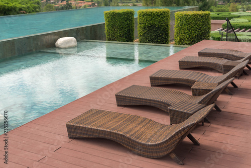 brown rattan sofa bed or bench, chair beside beautiful swimming pool, decorated with green bush of tree. home and resort decoration concept