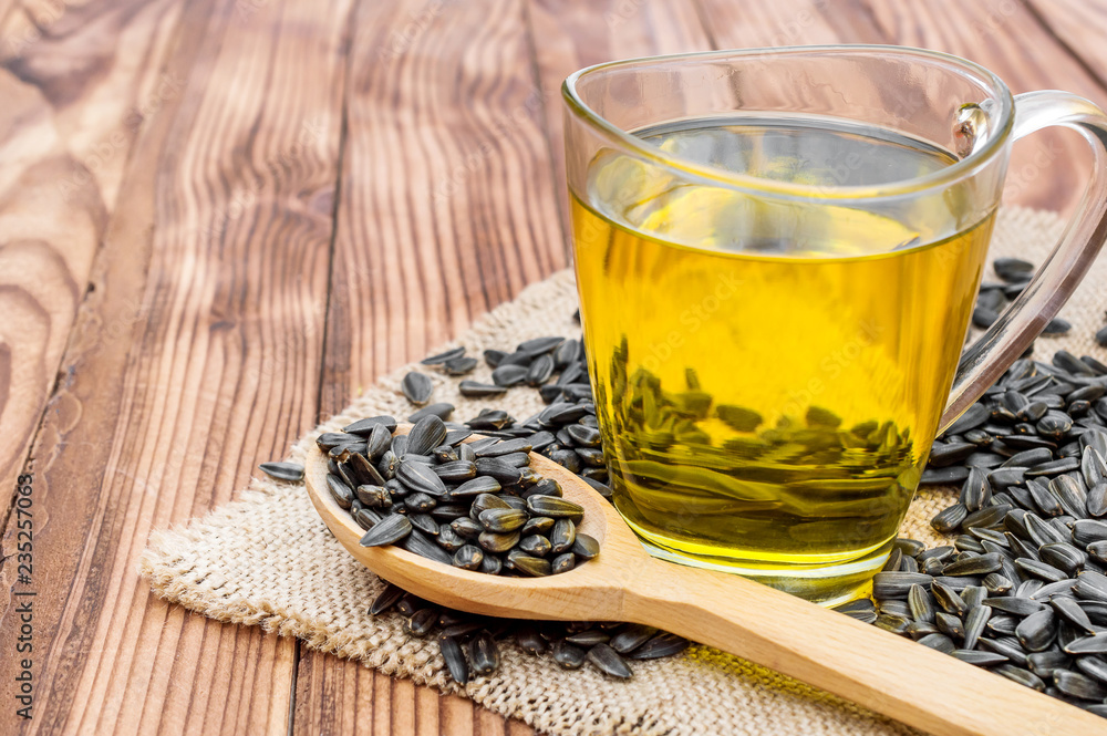 Fototapeta premium Glass cup of cooking oil and wooden spoon with sunflower seeds on napkin of burlap.