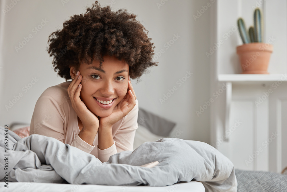 Lazy Afro American lady keeps both hands on cheeks, wears nightclothes, enjoys spare time in bed, has recreation in bedroom, broad smile. Housewife has rest from house routine. Morning concept Lazy Afro American lady keeps both hands on cheeks, wears nightclothes, enjoys spare time in bed, has recreation in bedroom, broad smile. Housewife has rest from house routine. Morning concept