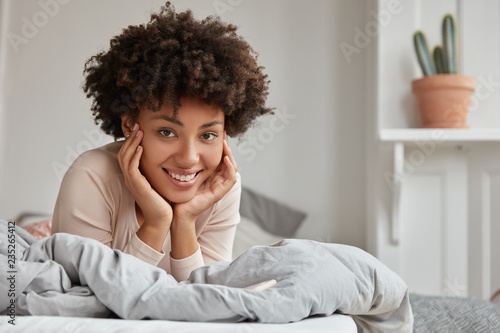 Lazy Afro American lady keeps both hands on cheeks, wears nightclothes, enjoys spare time in bed, has recreation in bedroom, broad smile. Housewife has rest from house routine. Morning concept