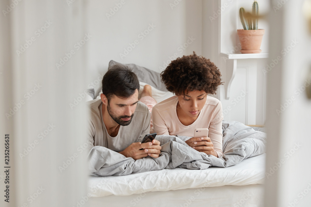 Image of mixed race woman and man wait for phone call, lie in comfortable bed, have rest at home, surf in social networks, have spare time during day off. Family, technology, leisure concept Image of mixed race woman and man wait for phone call, lie in comfortable bed, have rest at home, surf in social networks, have spare time during day off. Family, technology, leisure concept