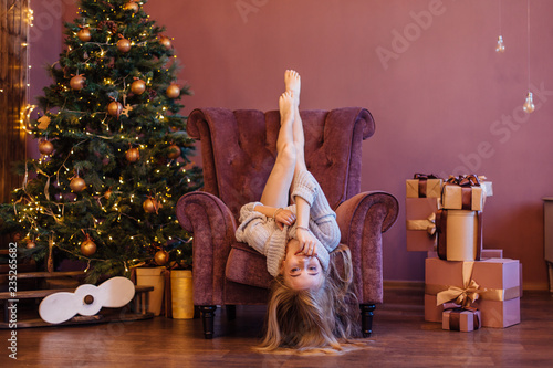 Beautiful young woman dressed in a sweater laying in the arm chair up side down next to the christmas tree.