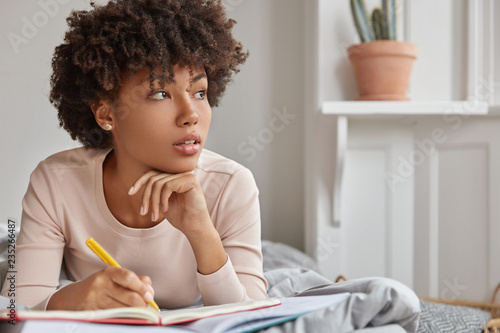 Canvas-taulu Close up shot of pensive dark skinned lady with Afro haircut, keeps hand under chin, notes text in notepad with yellow pen, wears casual clothing, lies in comfortable bed, focused aside