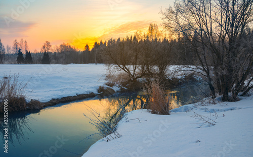 Frosty foggy winter landscape with small forest river and rising sun.