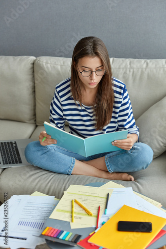 Indoor shot of woman freelancer focused into book, sits in lotus pose at comfortable sofa, surrounded with papers, works from home, records information from network, prepares for final exams