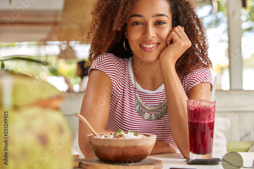 Photo of cheerful dark skinned girl smiles gladfully  eats delicious salad  drinks fresh cocktail  spends free time at coffee shop  wears casual clothing  has day off. People  lifestyle  ethnicity