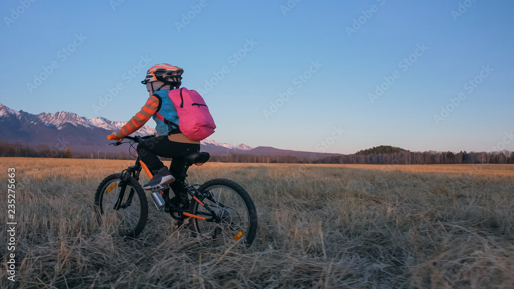 One caucasian children rides bike in wheat field. Little girl riding black orange cycle on background of beautiful snowy mountains. Biker motion ride with backpack and helmet. Mountain bike hardtail. One caucasian children rides bike in wheat field. Little girl riding black orange cycle on background of beautiful snowy mountains. Biker motion ride with backpack and helmet. Mountain bike hardtail.