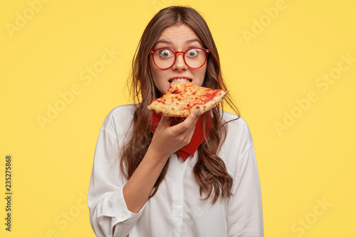 Emotive beautiful lady bites delicious pizza, looks directly at camera, has time for snack, visits pizzeria, surprised with low prices, models over yellow background. People, fast food and nutrition photo