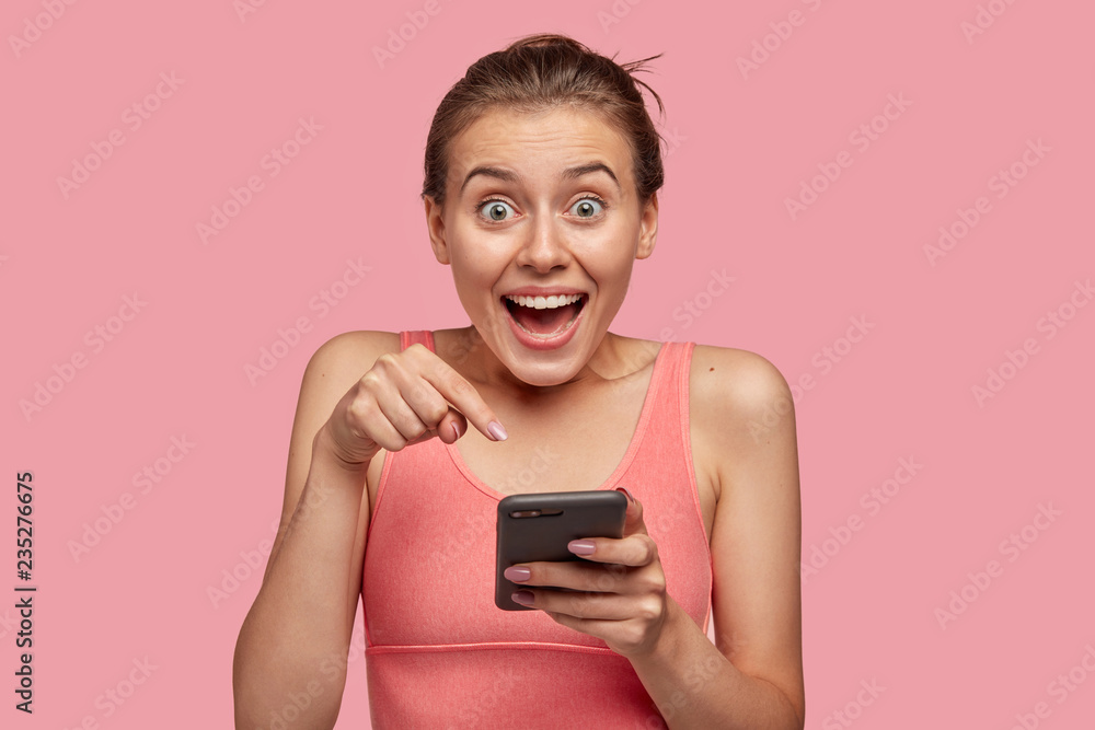 Photo of emotive Caucasian sporty woman has surprised expression, indicates with index finger in monitor of cellular, wears casual vest, recieves good notification, isolated over pink background Photo of emotive Caucasian sporty woman has surprised expression, indicates with index finger in monitor of cellular, wears casual vest, recieves good notification, isolated over pink background