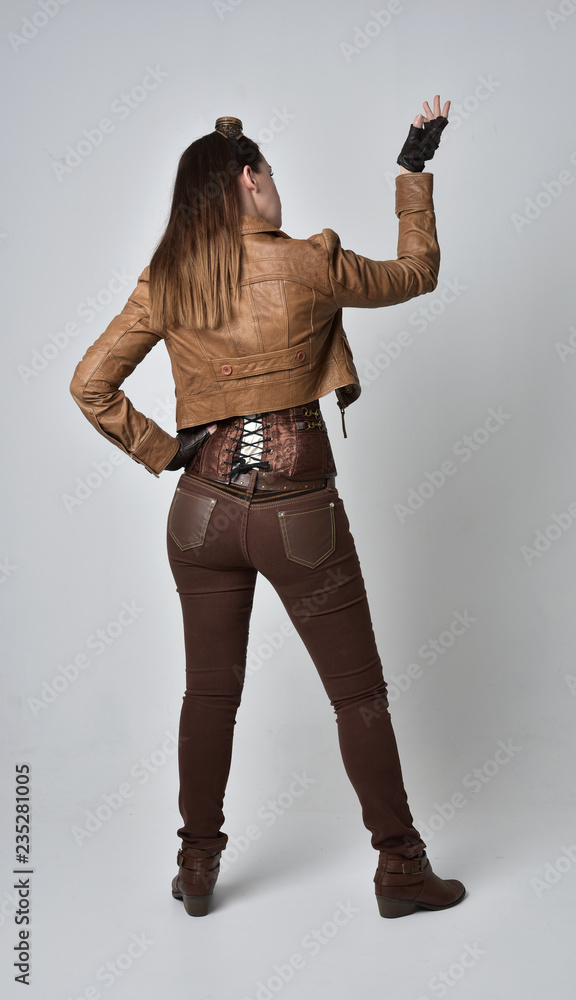 full length portrait of brunette girl wearing brown leather steampunk outfit. standing pose with back to the camera, on grey studio background. full length portrait of brunette girl wearing brown leather steampunk outfit. standing pose with back to the camera, on grey studio background.