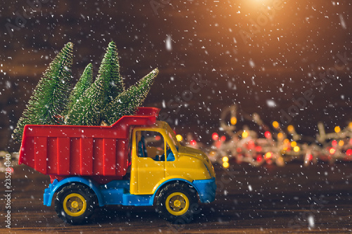 Little toy truck carrying christmas trees during snowfall. Beautiful background for greeting card. Winter composition. Happy holiday mood. Xmas and New Year fairytale wallpaper.