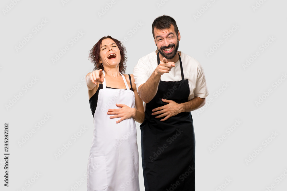 Couple of cooks pointing with finger at someone and laughing a lot on grey background Couple of cooks pointing with finger at someone and laughing a lot on grey background
