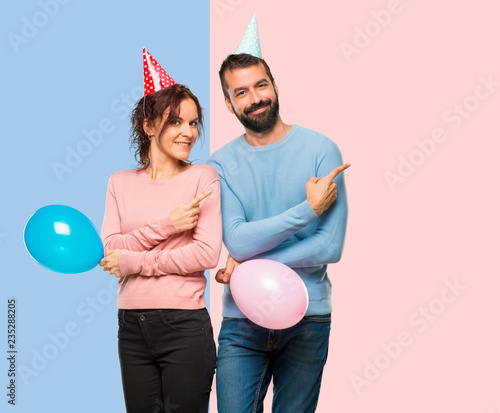 Wallpaper Mural couple with balloons and birthday hats pointing to the side with a finger to present a product on pink and blue background Torontodigital.ca
