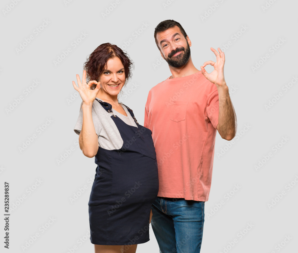 Couple with pregnant woman showing an ok sign with fingers on isolated grey background Couple with pregnant woman showing an ok sign with fingers on isolated grey background