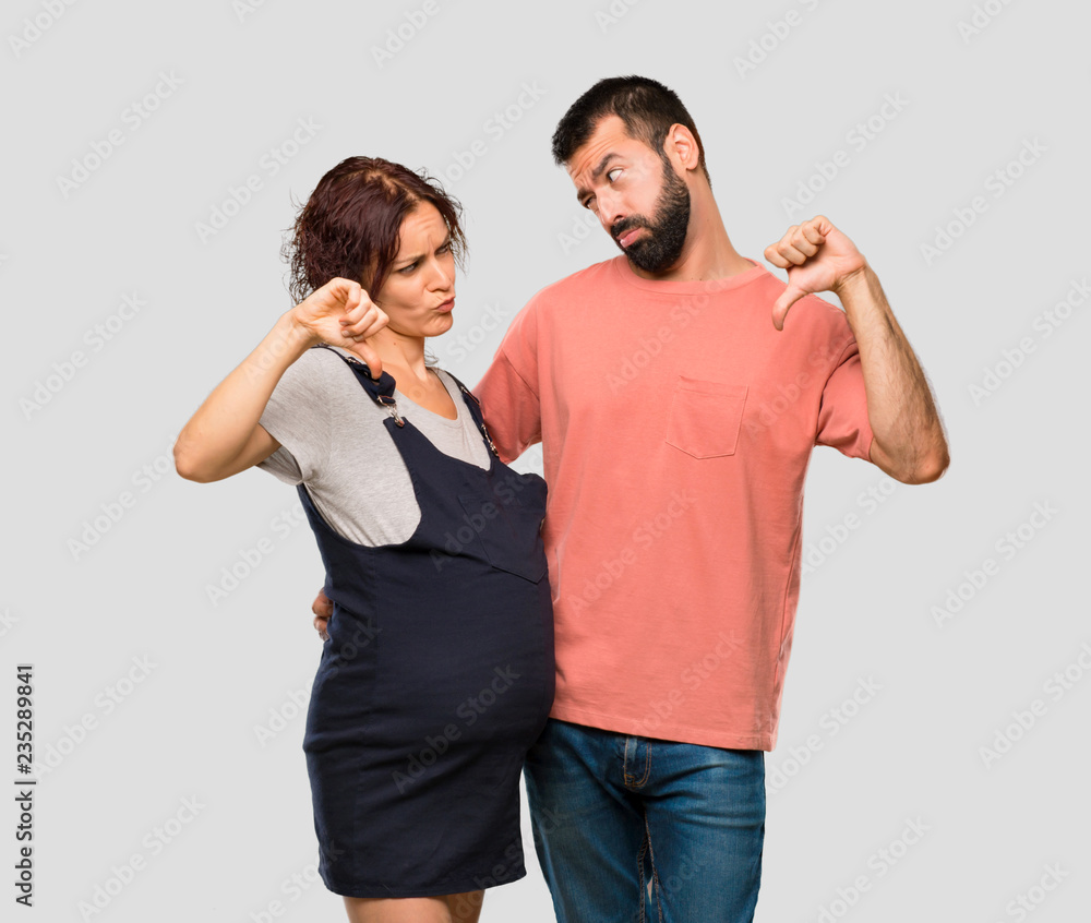 Couple with pregnant woman showing thumb down sign with negative expression on isolated grey background Couple with pregnant woman showing thumb down sign with negative expression on isolated grey background