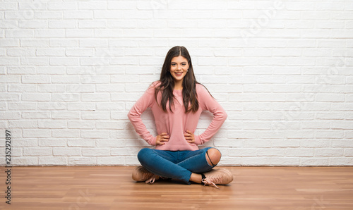 Teenager girl sitting on the floor in a room posing with arms at hip and laughing looking to the front