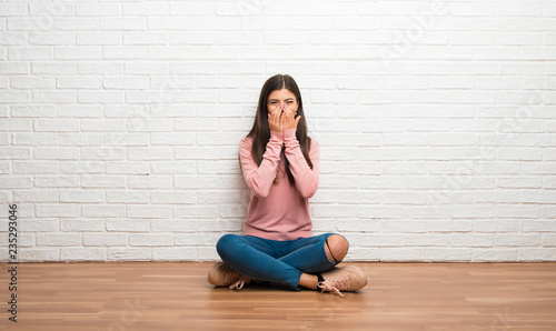 Teenager girl sitting on the floor in a room smiling a lot while covering mouth
