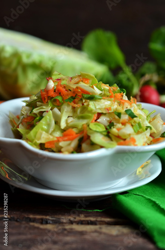 Young cabbage salad with carrot in a white bowl on a wooden background. Radishes and young cabbage in the background