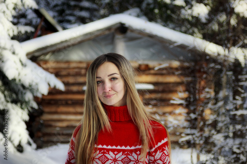 Portrait of a young woman in a red knitted sweater with a Norwegian pattern on the background of a hunting house in the winter snow-covered forest