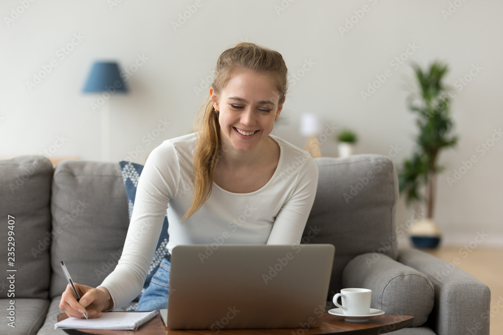 Smiling female sit on couch working on laptop, writing down important data,  happy girl have coffee studying online at computer at home, young woman  browse internet noting something relaxing on sofa Stock