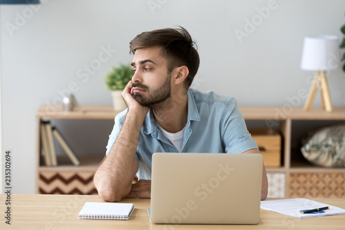 Tired male student or worker sit at home office desk look in distance having sleep deprivation, lazy millennial man distracted from work feel lazy lack motivation, thinking of dull monotonous job photo