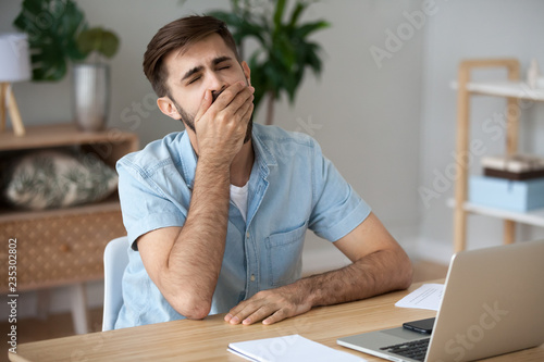 Slika na platnu Exhausted male worker sit at home office desk near laptop yawning working too lo