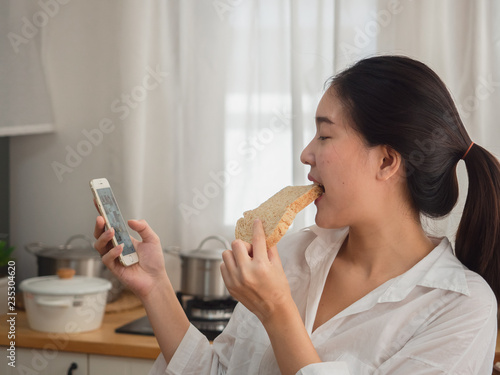 asian woman eating bread while using smart phone in the kitchen
