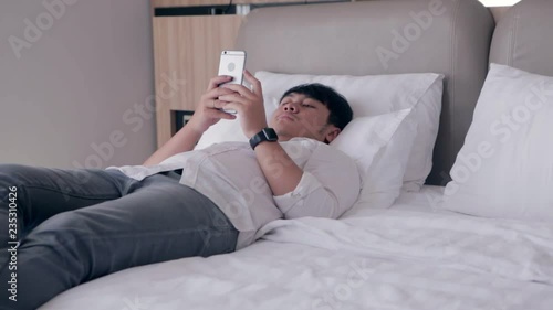 Young asian handsome man using smartphone laying down on bed while chatting with friend