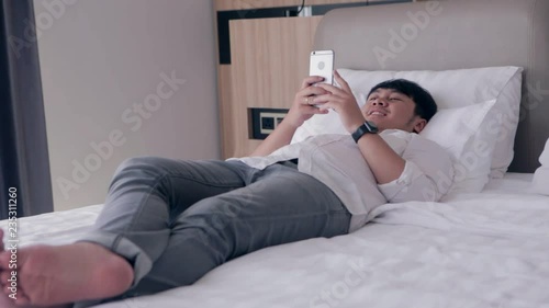 Young asian handsome man using smartphone laying down on bed while chatting with friend