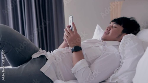 Young asian handsome man using smartphone laying down on bed while chatting with friend