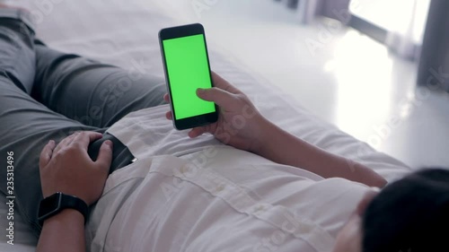 Back view of young asian handsome man using smartphone with green screen laying down on bed while chatting with friend