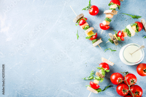 Healthy summer snack idea, salad sandwiches kebab on skewers, with bread slices, tomato, greens, cucumber, cheese, sausages, with yoghurt dipping, on light concrete background copy space top view