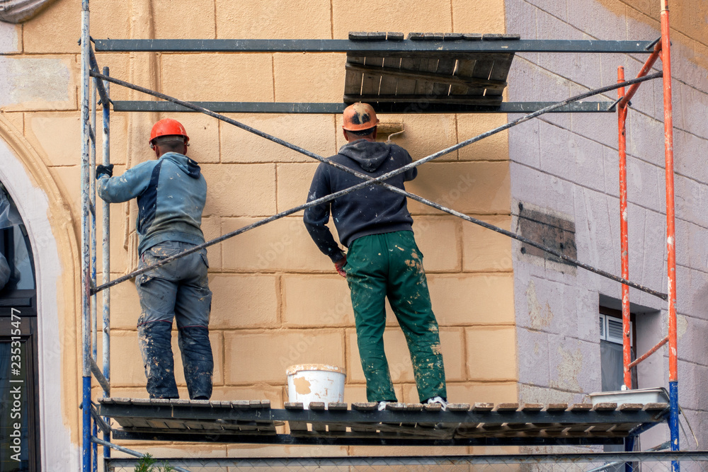 two workers in a helmet and construction clothes on a high staircase are painting with rollers, repairing the facade of the house two workers in a helmet and construction clothes on a high staircase are painting with rollers, repairing the facade of the house