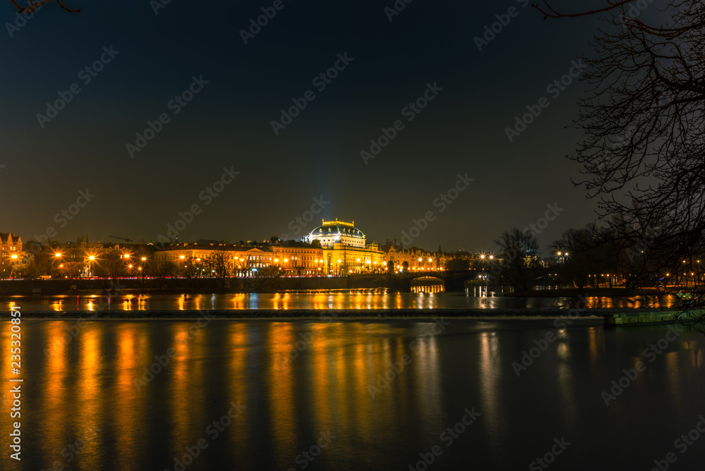 Illuminated classical buildings and street lights reflecting on the Vltava river in Prague before a small waterfall in a cold winter night - 1