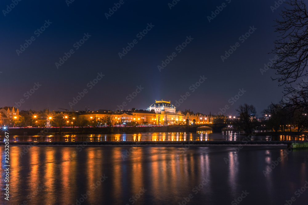 Illuminated classical buildings and street lights reflecting on the Vltava river in Prague before a small waterfall in a cold winter night - 2