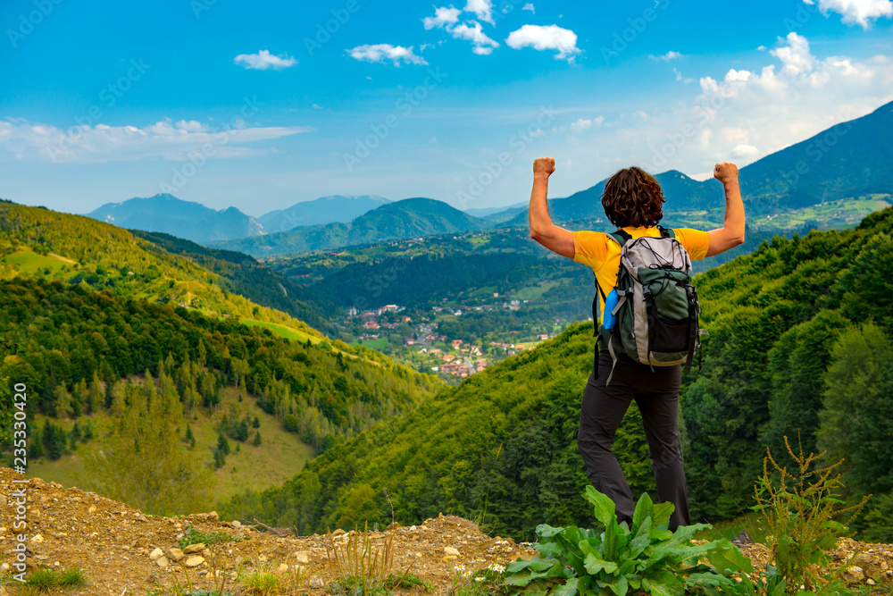 Young mountain hiker with arms raised up, celelbrating and enjoying a beautiful mountain landscape covered with lush forests. Hiking in a sunny summer day.