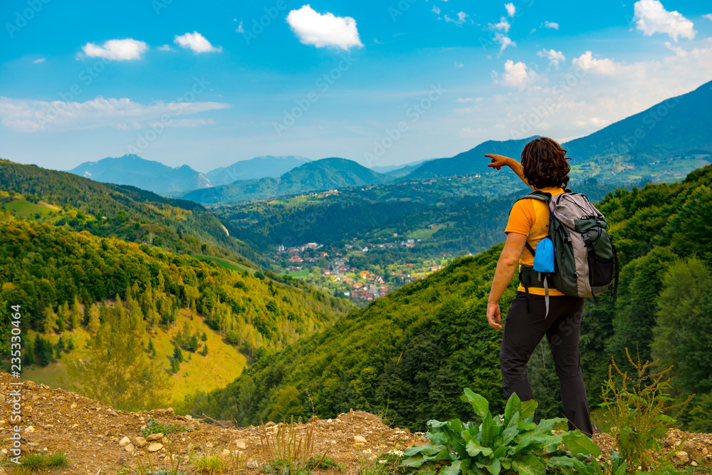 Young mountain hiker pointing to a beautiful mountain landscape covered with lush forests. Hiking in a sunny summer day.