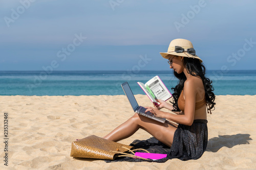 Asian sexy woman in bikini, using laptop computer and holding book on a beach, travel of summer vacation. Freelance work concept.