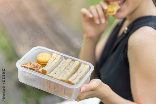 Woman holding appetizer lunch box  and eating healthy peanut butter sandwich cracker sitting at park.